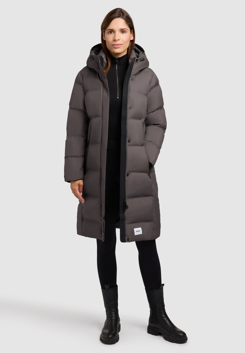 Padded gray coat with a hood, closed with buttons, featuring a logo patch at the hem, paired with black pants and knee-high boots.
