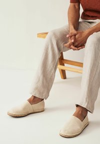 Beige canvas espadrilles with a natural jute sole, featuring a slip-on design and rounded toe, set against light-colored trousers and a wooden bench.