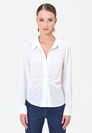 Donna con i capelli pettinati all'indietro indossa una camicia bianca arricciata con bottoni e jeans blu scuro, in piedi su uno sfondo chiaro e uniforme.