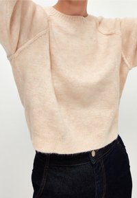 Femme portant un pull en maille couleur pêche et un jean bleu foncé, levant les bras, sur un fond clair uni.