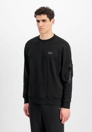 Joven con sudadera negra de manga larga con bolsillo con cremallera y detalle de anilla en la manga izquierda, de pie frente a un fondo blanco liso.