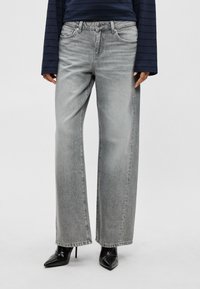 Asmuo dėvi pilkus plačių kojų džinsus, juodus aukštakulnius batus su smailiais pirštais ir tamsiai mėlyną dryžuotą ilgomis rankovėmis palaidinę.