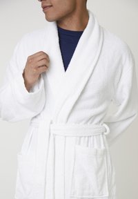 Homme portant un peignoir en éponge blanc par-dessus une chemise bleu foncé, tenant le col du peignoir d'une main.