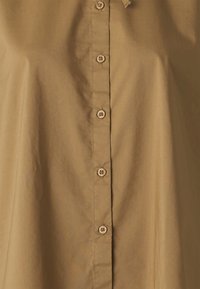 Chemise beige à manches longues avec une texture lisse, dotée d'une rangée de boutons ronds de couleur claire sur le devant et d'une coupe décontractée.