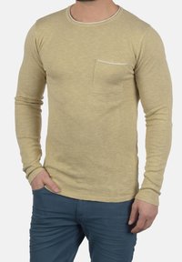 Pitkähihainen paita vaaleanbeigessä, valmistettu kuvioidusta kankaasta. Varustettu pyöreällä pääntiellä ja edessä olevalla taskulla. Yhdistetty sinisiin housuihin.