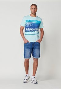 Ljusblå grafisk t-shirt med palmbladdesign och horisontella ränder, kombinerad med uppvikta denimshorts och vita sneakers.