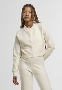 Krémově zbarvený krátký kardigan s knoflíkovým zapínáním vpředu, žebrovanými lemy na rukávech a límcem. Vyrobený z měkké tkaniny; nošený s odpovídajícími kalhotami.