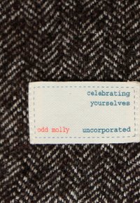 Vlněná tkanina se vzorem rybí kosti, převážně tmavě hnědá s nádechy krémové barvy. Bílé štítek s červeným a modrým textem nese nápis "odd molly."