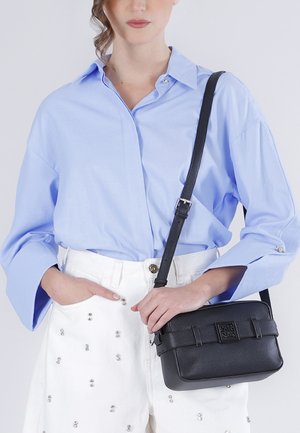Kobieta ubrana w jasnoniebieską, oversize'ową koszulę i białe spodnie z srebrnymi ćwiekami, trzymająca czarną skórzaną torebkę na ramię z przednią klamrą.