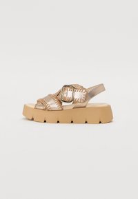 Sandale avec une plateforme épaisse beige, des brides métalliques dorées et une boucle décorative. Présente des accents texturés le long des bords des brides.