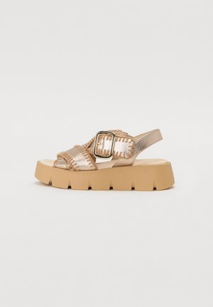 Sandale avec une plateforme épaisse beige, des brides métalliques dorées et une boucle décorative. Présente des accents texturés le long des bords des brides.