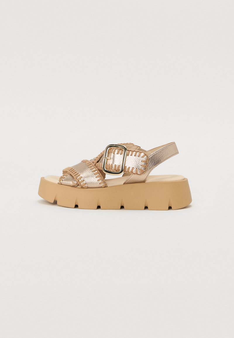 Sandale avec une plateforme épaisse beige, des brides métalliques dorées et une boucle décorative. Présente des accents texturés le long des bords des brides.