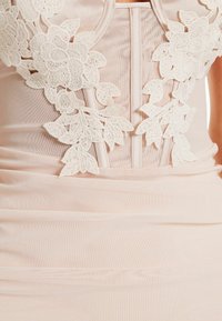 Robe rose pâle ornée d'un appliqué floral en dentelle blanche délicate, avec un corsage structuré et une texture de tissu froncé, soulignant une silhouette ajustée.