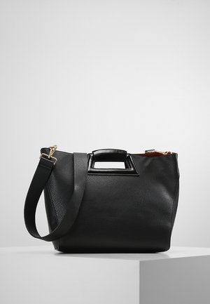 Black textured leather handbag with rectangular cutout handle, detachable shoulder strap, and gold-tone hardware on a white pedestal.