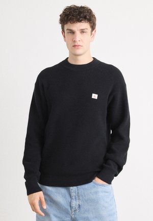 Svart strikket genser med rund hals og ribbestrikket tekstur. Har et lite logo-patch på brystet. Kombinert med lyseblå denimjeans.