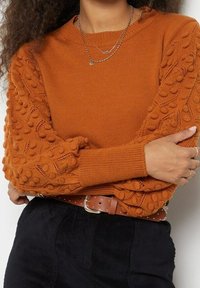 Pull en tricot couleur rouille avec manches texturées et ornées, poignets côtelés, associé à une ceinture marron ornée de clous, sur un vêtement foncé.
