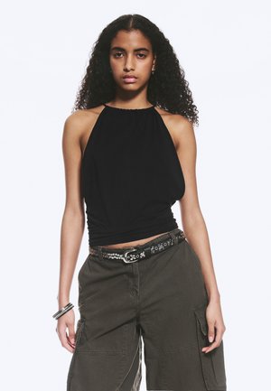 Jeune femme aux cheveux bouclés portant un top dos nu noir, un pantalon cargo foncé avec une ceinture cloutée et des bracelets argentés au poignet gauche.
