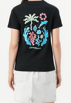 Sort t-shirt med farverigt havtema-grafik, der viser koraller, fisk, bølger og en palme, båret med en hvid nederdel.