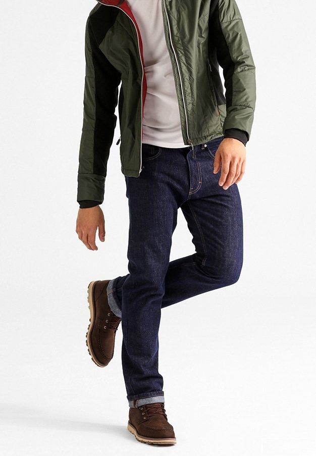 Homme portant un jean bleu foncé, des bottes marron, une chemise blanche et une veste verte avec une doublure rouge, se tenant sur une jambe sur un fond blanc.