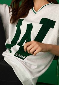 Biała bawełniana bluza z dekoltem w serek, z zielonym nadrukiem "Wicked" oraz dużą zieloną literą "W", charakteryzująca się fakturowaną powierzchnią i kontrastującym prążkowanym wykończeniem przy szyi.