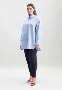 Camicia oversize a righe blu e bianche con colletto classico con bottoni, maniche lunghe e orlo curvo, abbinata a pantaloni neri affusolati e sandali con cinturini.