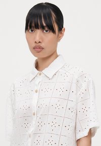 Camisa blanca de manga corta con tejido de broderie anglaise y patrones florales, cuello pico y cierre frontal con botones.