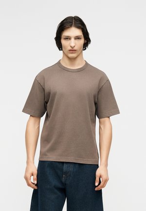 Νέος με κοντά, σκούρα μαλλιά, που φοράει ένα απλό καφέ κοντομάνικο μπλουζάκι και φαρδιά σκούρα μπλε τζιν, μπροστά σε λευκό φόντο.