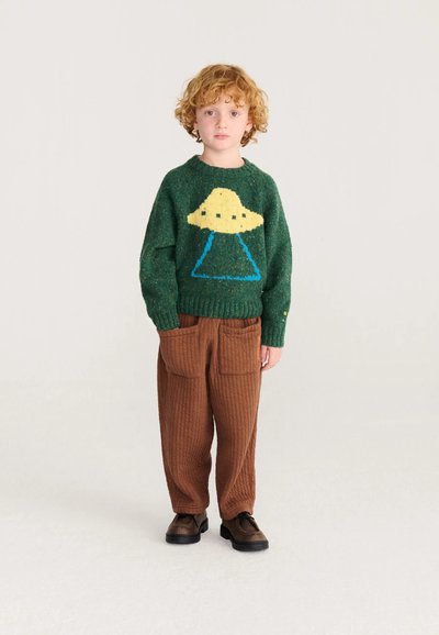 Enfant aux cheveux bouclés portant un pull vert avec un motif UFO jaune, pantalon matelassé marron avec de grandes poches et chaussures marron, debout.