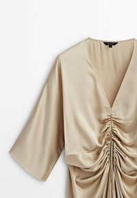 Blouse en satin beige avec un profond décolleté en V, manches larges, et détail froncé au centre devant avec des cordons ajustables.