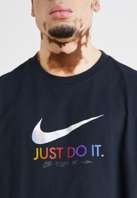 Juodas medvilnės marškinėliai su dideliu švytinčiu Nike logotipu ir daugiakolorės „JUST DO IT“ tekstu priekyje. Paprastos apvalios kaklo dizainas.