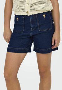 Pantalones cortos de mezclilla en azul oscuro con un acabado texturizado. Presentan dos bolsillos delanteros, detalles metálicos dorados y un dobladillo con vuelta.
