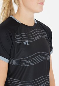 Musta urheilupaita harmaalla aallokkuvalla kuviolla ja vaaleanharmailla yksityiskohdilla kauluksessa ja hihoissa. Hengittävää, joustavaa kangasta. FZ-logo rinnassa.