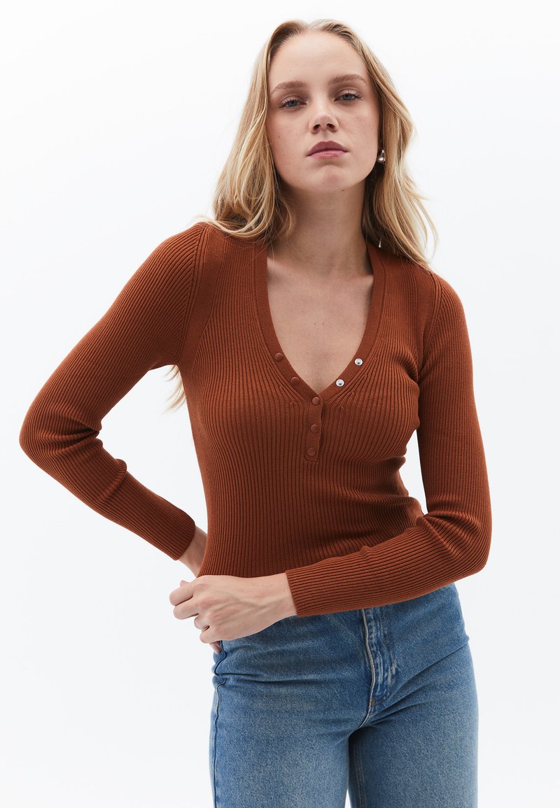 Top de manga larga acanalado en color óxido con un profundo escote en V y tres botones. Combinado con unos jeans de mezclilla azul de cintura alta.