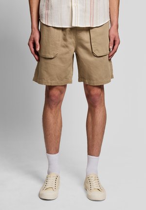 Person, die beige knielange Cargoshorts, weiße Socken und cremefarbene Sneakers trägt, vor einem einfarbigen grauen Hintergrund stehend.