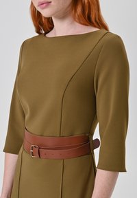 Olijfkleurige jurk met een boothals en driekwart mouwen, voorzien van een bruine leren riem met een gesp en gestikte details.