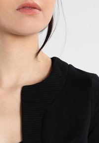 Musta vaate, jossa on pyöreä pääntie ja ribbauskaulus. Silkkinen kangas, ilman näkyviä kuvioita tai metallidetaljeja.