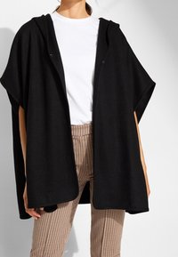 Kobieta ubrana w luźny czarny płaszcz z kapturem, na białą koszulkę i w kratkę spodnie, stojąca na tle jasnego, jednolitego tła.