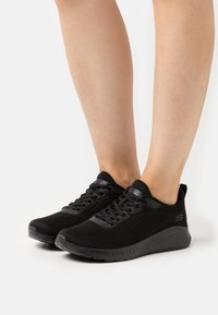 Black athletic shoes with mesh upper, textured fabric, padded collar, and rubber sole. Features laces and logo on the side.