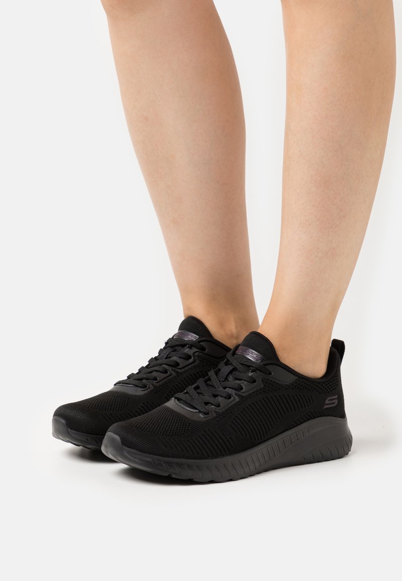 Black athletic shoes with mesh upper, textured fabric, padded collar, and rubber sole. Features laces and logo on the side.