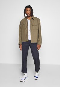 Olivenfarvet uldskjortejakke med to brystlommer, parret med en hvid t-shirt, marineblå bukser optrukket ved anklerne, og multikolor sneakers.