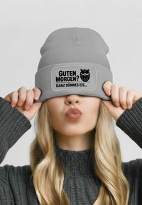 Gray beanie hat with a flat brim and a rectangular patch. Patch features black text and an owl graphic. Model poses with hat covering eyes.