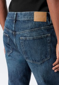 Blugi din denim albastru cu un finisaj texturat, având două buzunare la spate și o etichetă din piele maro, inscripționată "WEEKDAY" pe talie.