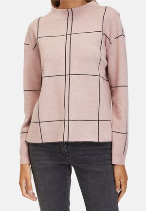 Vrouw draagt een lichtroze trui met grote zwarte rasterlijnen, gecombineerd met donkergrijze jeans, staand tegen een witte achtergrond.