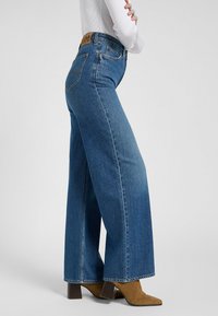 Jeans in denim blu a gamba larga con vita alta, dotati di un'etichetta in pelle sul retro e una leggera texturesfocata. Stivaletti con tacco marroni completano il look.