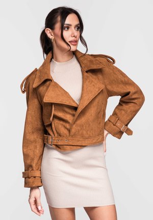 Femme portant une veste courte en daim marron sur une robe beige en tricot côtelé, posant avec une main sur la hanche et regardant de côté.