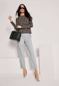 Vrouw in houndstooth trui, lichtgrijze broeken, zilveren hakken, grote zwarte zonnebril, met een zwarte handtas, staande tegen een effen achtergrond.