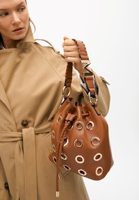 Brown leather bucket bag with gold grommets, braided drawstring closure, and a patterned strap featuring golden accents.