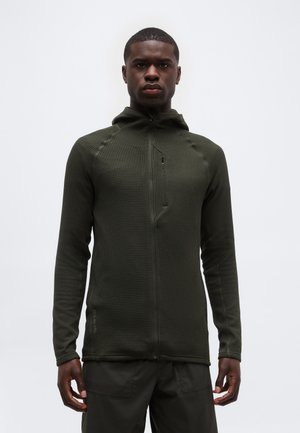 Homme portant un hoodie zippé texturé vert foncé et un pantalon assorti, debout devant un fond clair uni, faisant face à l'avant.