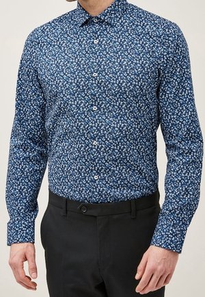 Homme portant une chemise bleue ajustée à manches longues avec des motifs floraux blancs, boutonnée à l'avant, associée à un pantalon noir.