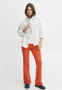 Hvid denimjakke med sølvknapper over en hvid skjorte, parret med lyseorange flagrende bukser og sorte kraftige støvler. Simpelt design.